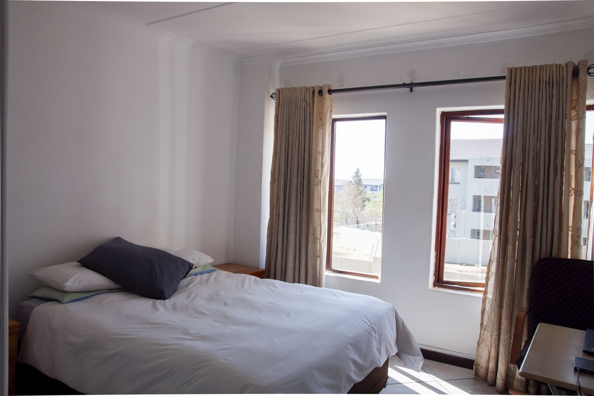A functional bedroom featuring tiled flooring and dual casement windows with installed curtain rails, offering natural light and a view of the surrounding complex.