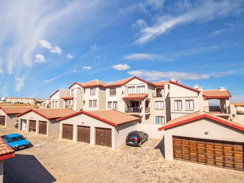 Exterior view of a multi-story apartment complex featuring a paved driveway, multiple garage units, and tiled roofing under a clear blue sky.