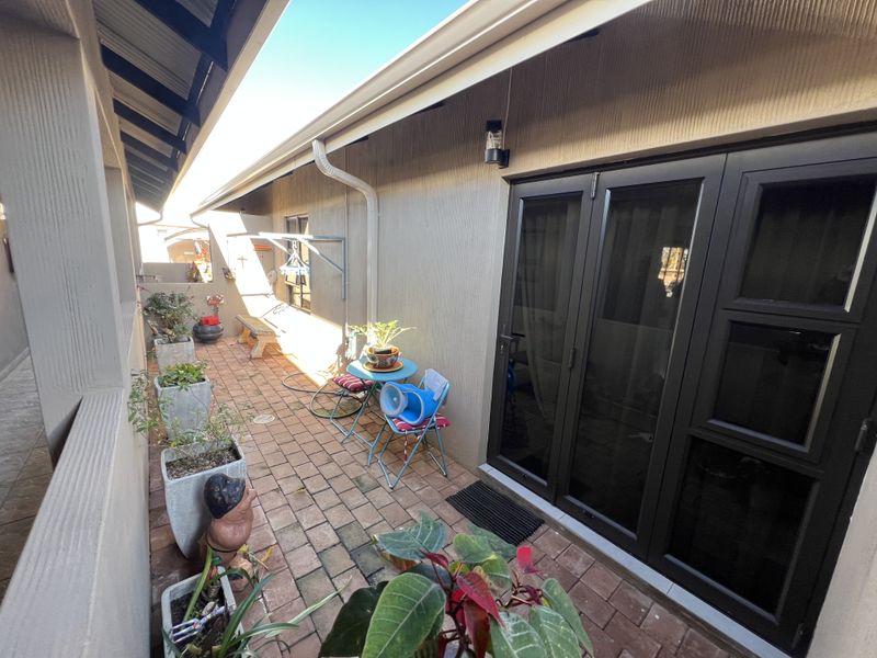 A covered patio area featuring brick paving, wall-mounted lighting, and dark-framed glass sliding doors providing access to the interior.