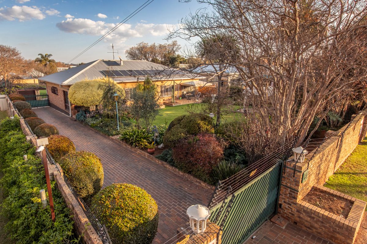 Aerial view of the property featuring a brick-paved driveway, landscaped garden, solar panels on the roof, and secure perimeter walling with electric fencing.