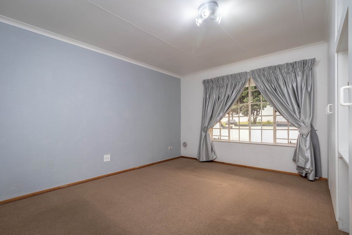 A spacious, empty bedroom featuring neutral carpet flooring, a built-in cupboard, and a large window with security bars.
