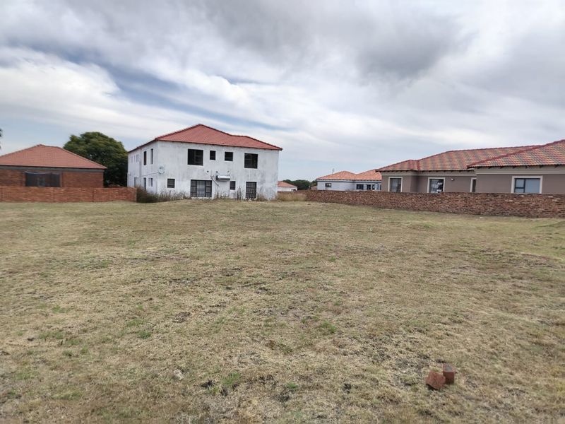 A view of a vacant residential plot featuring a brick boundary wall and surrounding residential properties with tiled roofs.