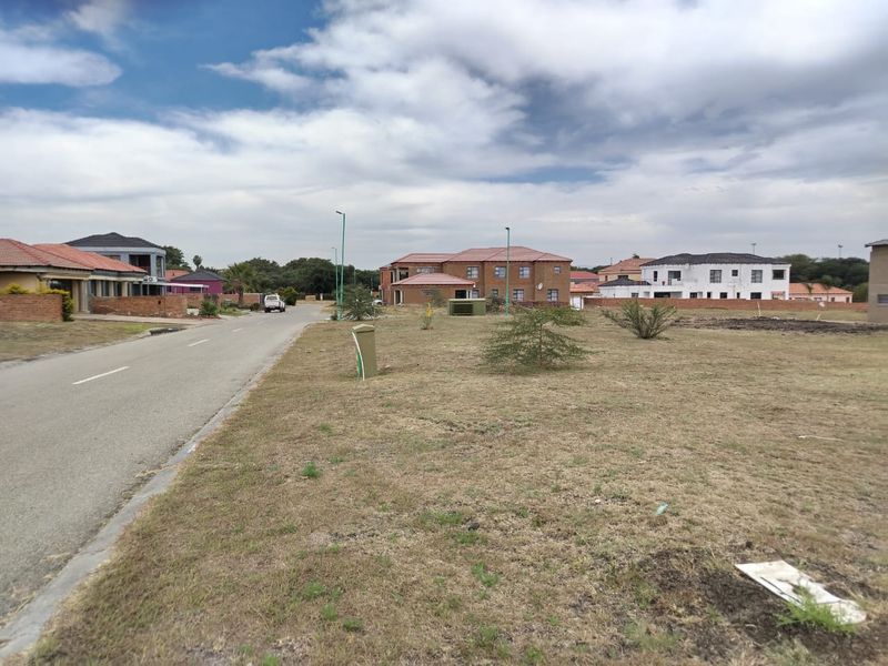 A street-level view of a vacant residential plot in a developed neighborhood, featuring paved road access and existing utility connections.