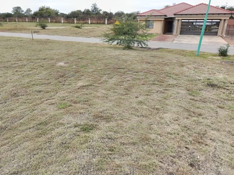 A view of a vacant residential plot featuring a grass lawn, adjacent to a paved road and existing residential development.