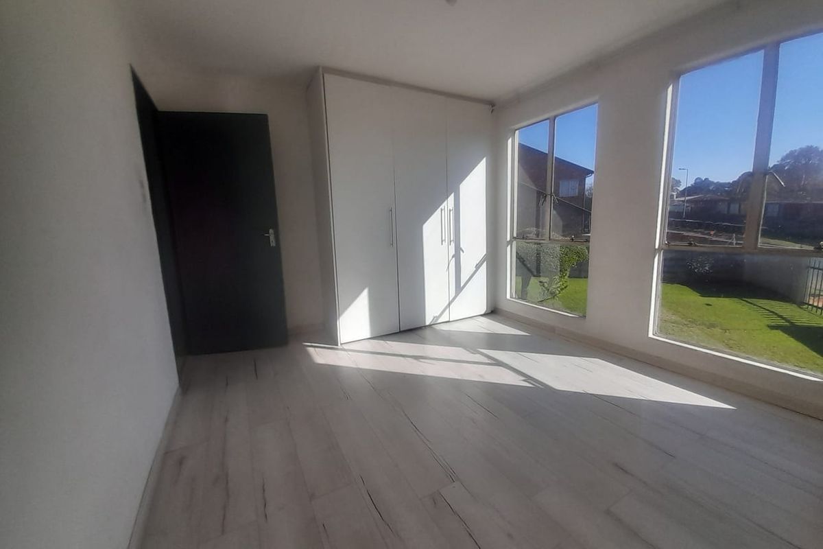 A bright, empty bedroom featuring light-toned laminate flooring, a built-in wardrobe, and large windows providing ample natural light.