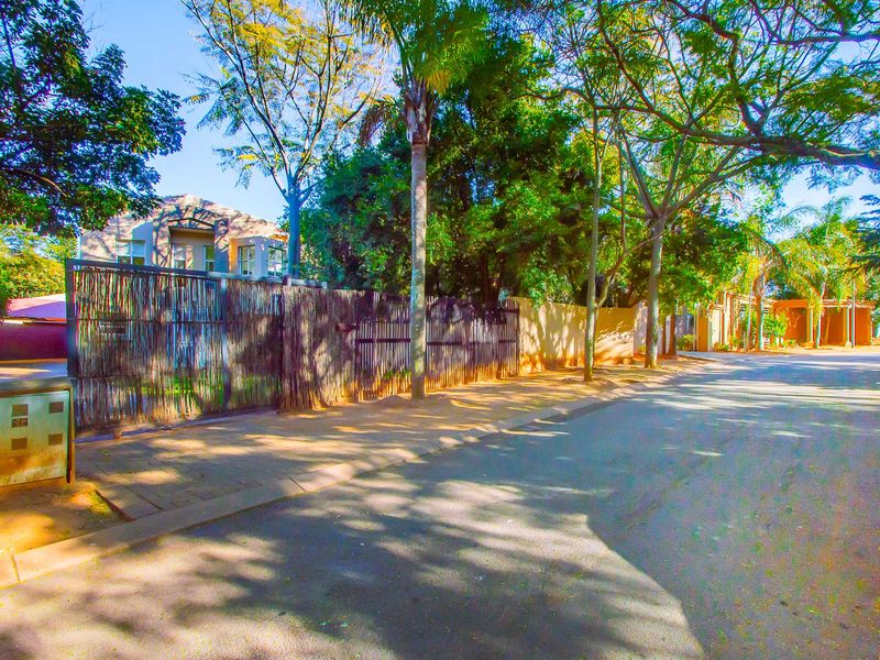 A street-level view of the property frontage, featuring mature trees, a paved sidewalk, and a boundary fence along the asphalt road.