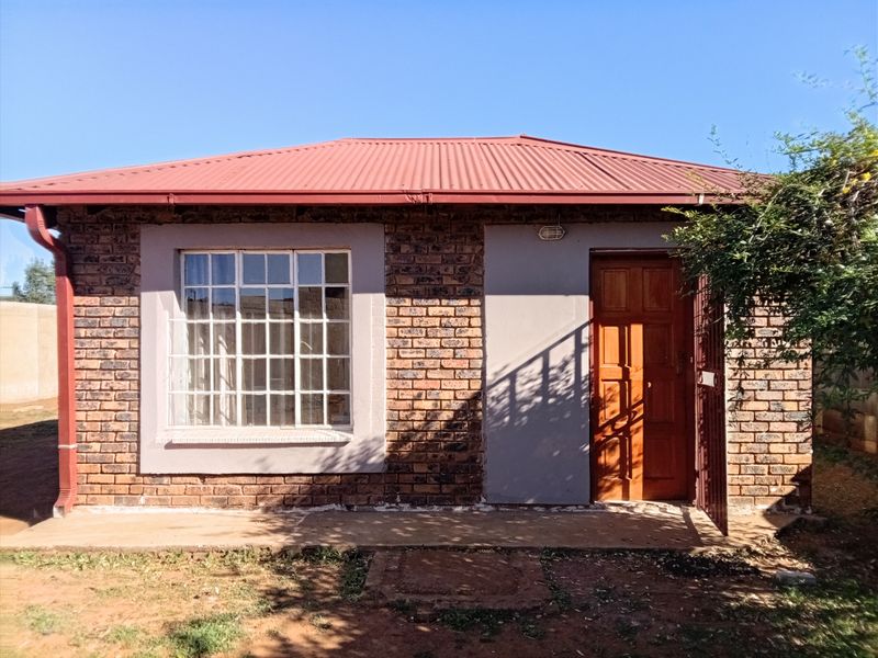 A front exterior view of a single-story facebrick house featuring a red corrugated metal roof, timber entrance door, and steel-framed windows.