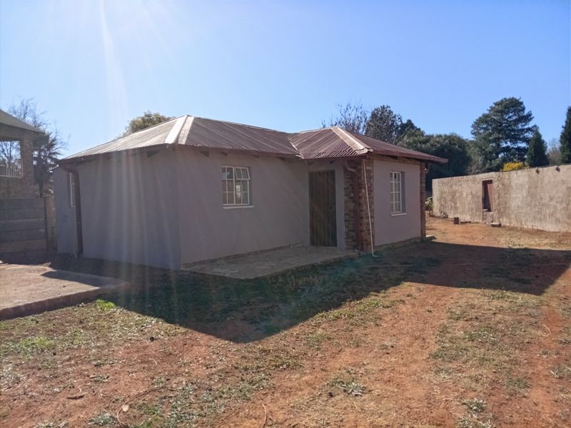 A single-story residential house featuring a corrugated metal roof, plastered walls, and a stone-paved entrance area set on a large plot.