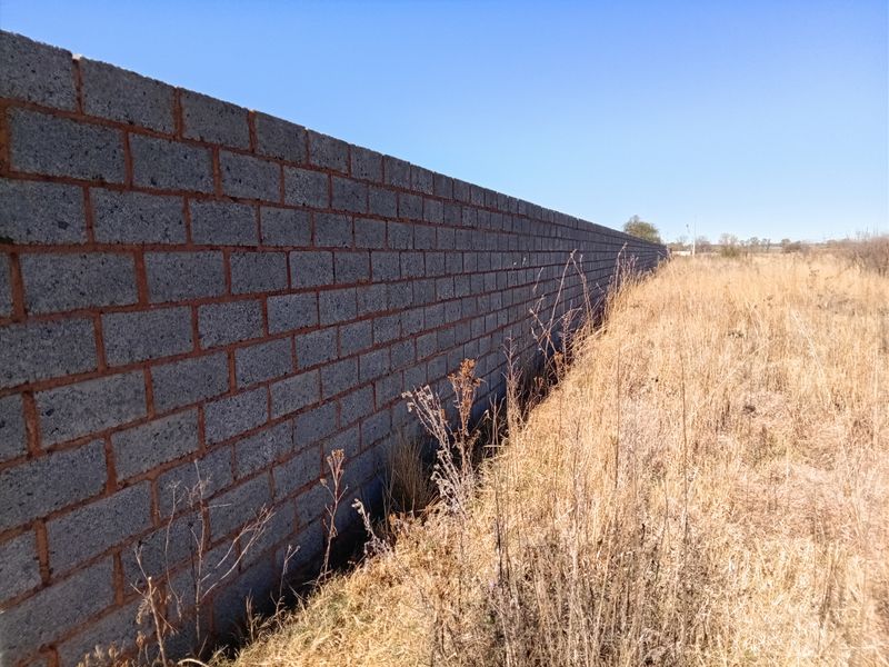 A long concrete block boundary wall bordering a vacant residential plot with dry grass.