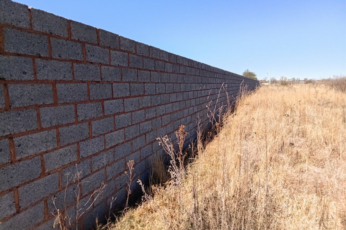 A long concrete block boundary wall bordering a vacant residential plot with dry grass.
