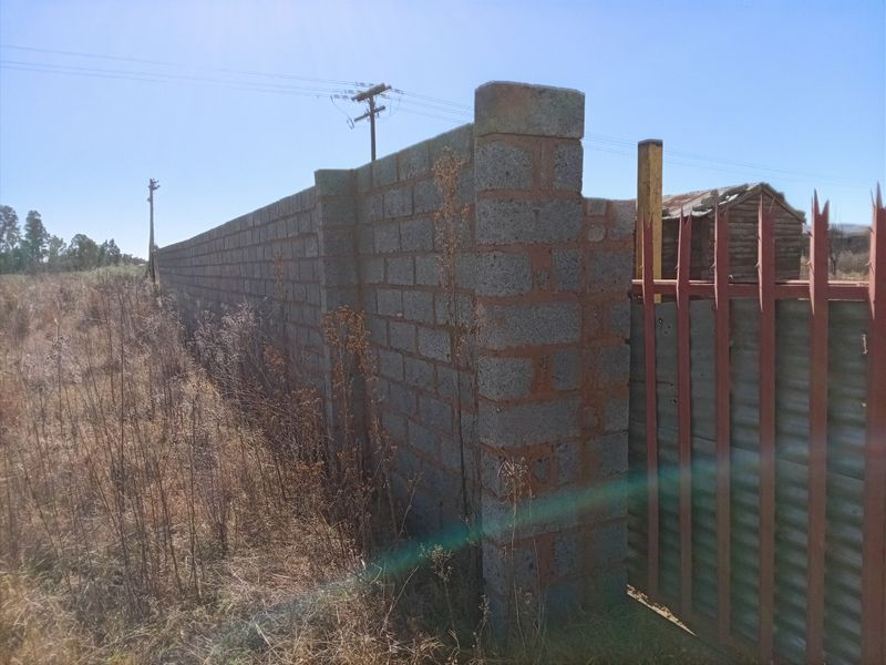 A view of a vacant residential plot featuring a concrete block boundary wall and a corrugated metal gate with security spikes.