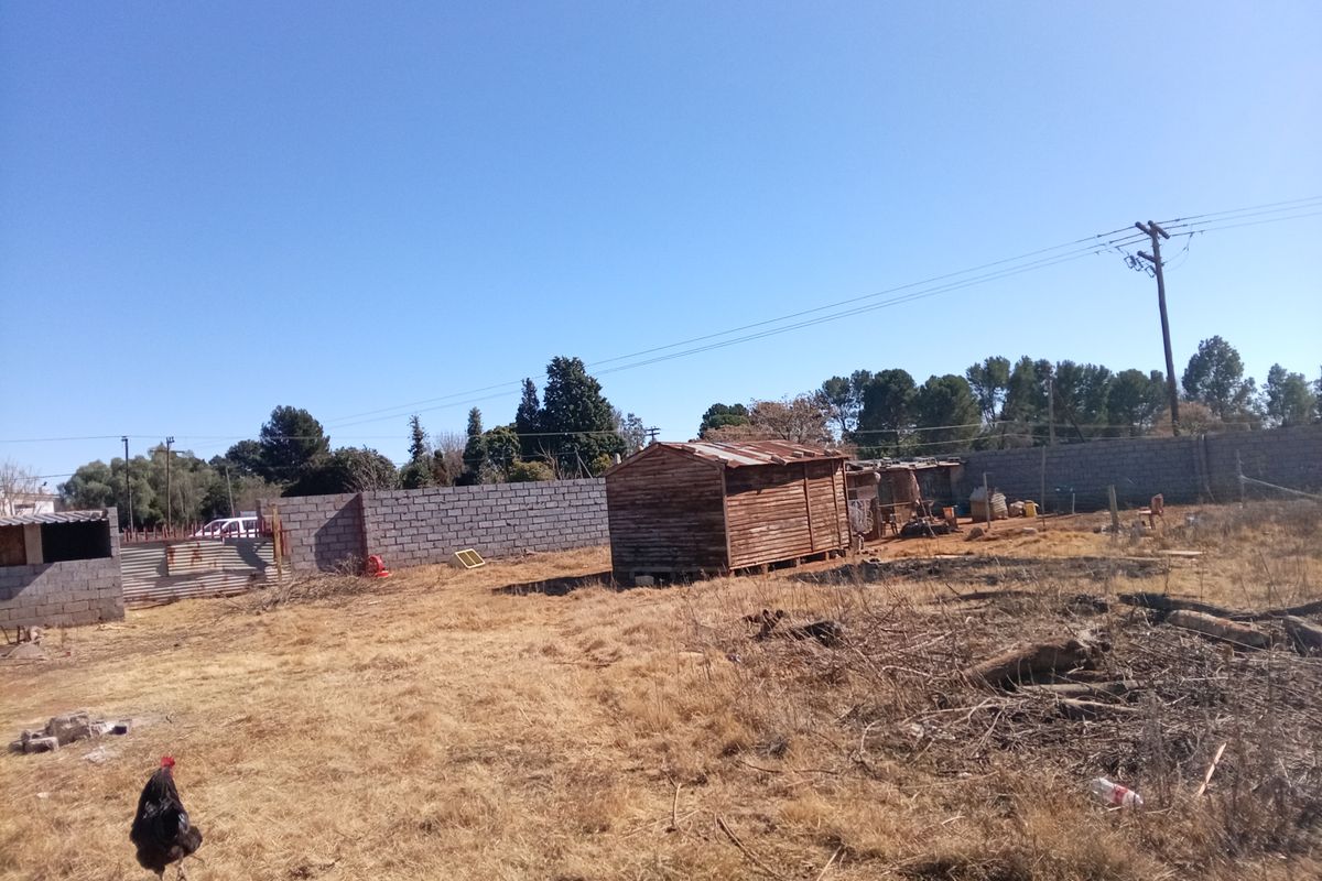 A vacant residential plot featuring a perimeter masonry wall, a small wooden shed, and dry, unmaintained ground cover.