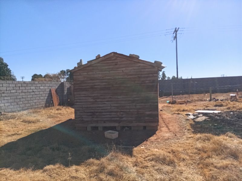 A view of a small timber shed structure situated on a vacant residential plot, enclosed by a concrete block boundary wall.