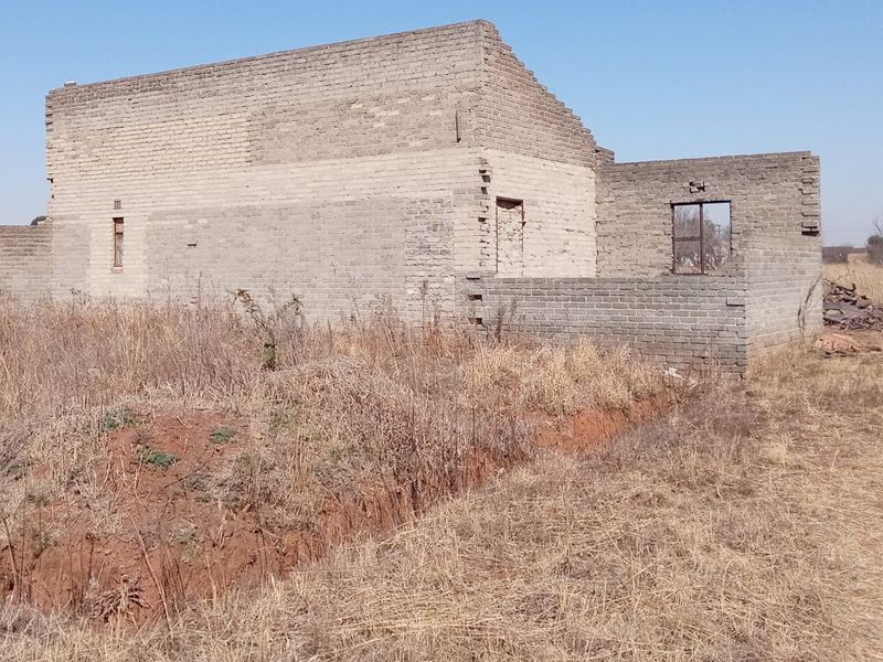 An incomplete brick structure showing the exterior shell of a building under construction, set on a vacant plot of land.