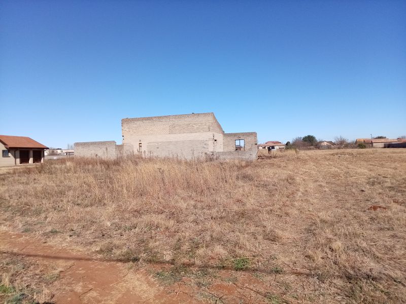 An unfinished concrete block structure set on a large, undeveloped plot of land, showing the early stages of residential construction.