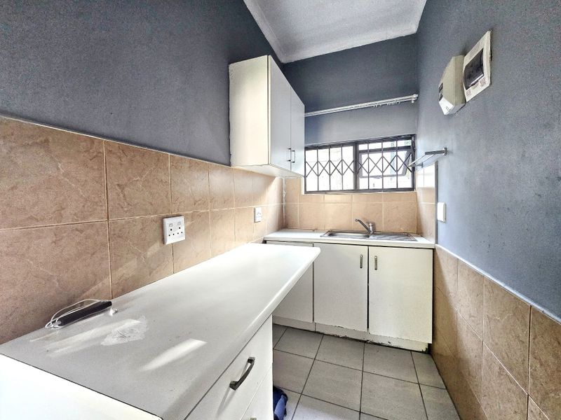 A functional scullery or utility area featuring built-in cabinetry, a stainless steel sink, tiled splashbacks, and secure window bars.