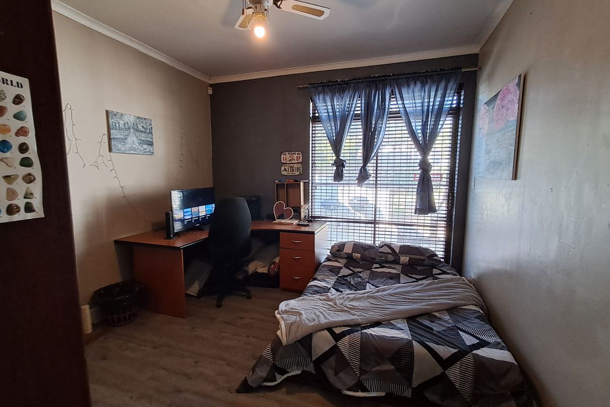 A functional bedroom featuring laminate flooring, a ceiling fan, and large windows with venetian blinds, offering a practical space for rest or study.