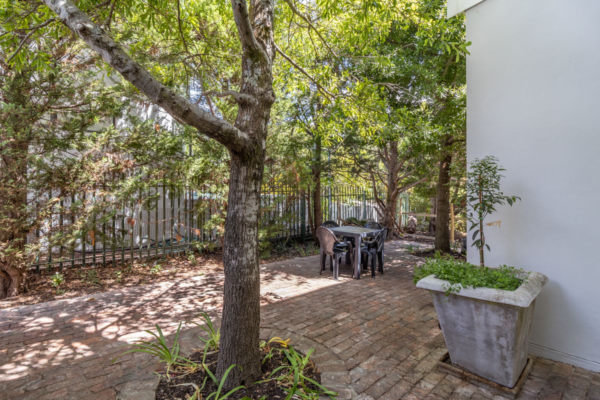A private rear garden area featuring brick paving, mature trees, and secure metal perimeter fencing, offering a quiet outdoor space.