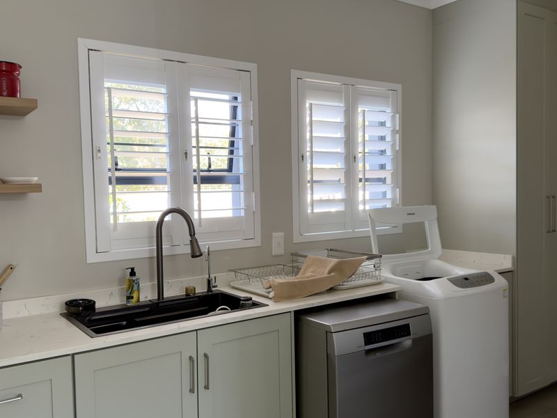 A well-appointed laundry room featuring sage green cabinetry, quartz countertops, an undermount sink with a modern faucet, and plantation shutters.