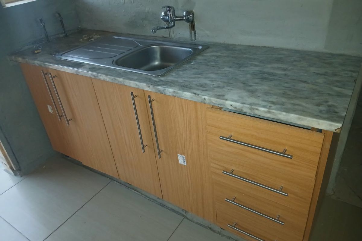 A kitchen area featuring a stainless steel sink, laminate countertop, and wood-finish cabinetry, currently in an unfinished state with exposed wall plaster and plumbing.