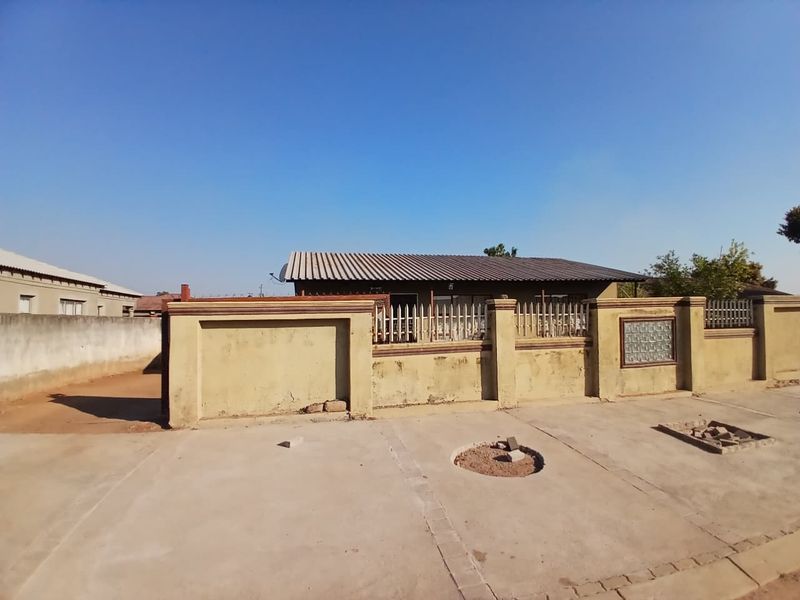 Front exterior view of a residential property featuring a concrete driveway, perimeter boundary wall with metal security fencing, and a corrugated metal roof.