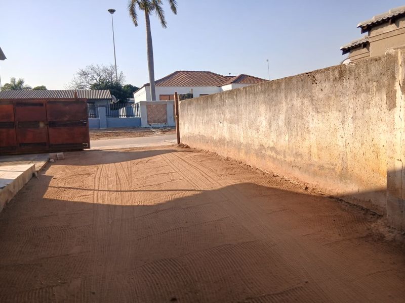 An exterior view of the property entrance featuring a metal gate and a long boundary wall with an unpaved driveway area.