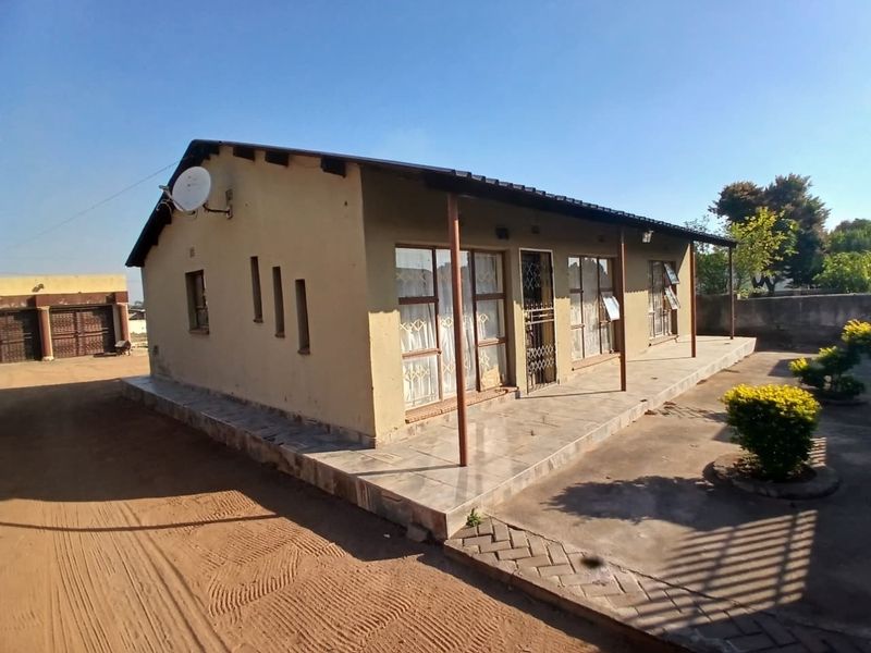Single-story residential house featuring a tiled front veranda, metal security entrance, and a metal roof, set on a large plot with a concrete driveway.