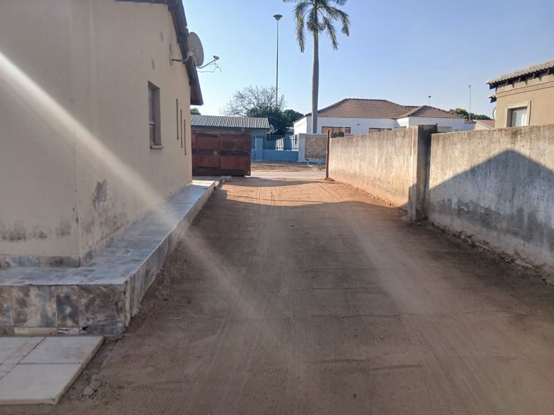 A view of the side or rear exterior area featuring a sandy driveway, boundary wall, and tiled plinth along the building facade.