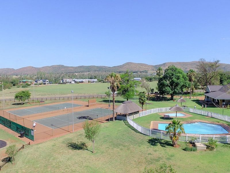 Aerial view of a residential estate featuring a tennis court, swimming pool with thatched lapa, and expansive landscaped grounds.