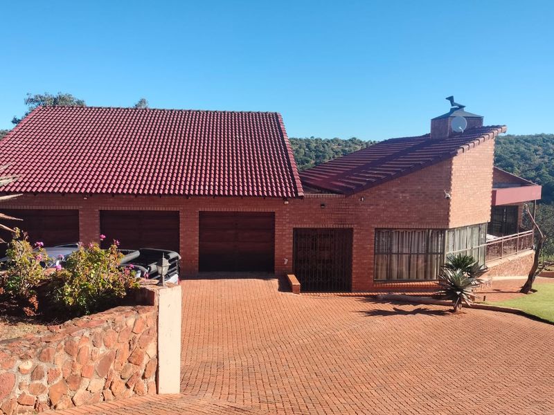 A well-maintained brick house featuring a triple garage, tiled roof, and an expansive paved driveway set against a natural landscape.