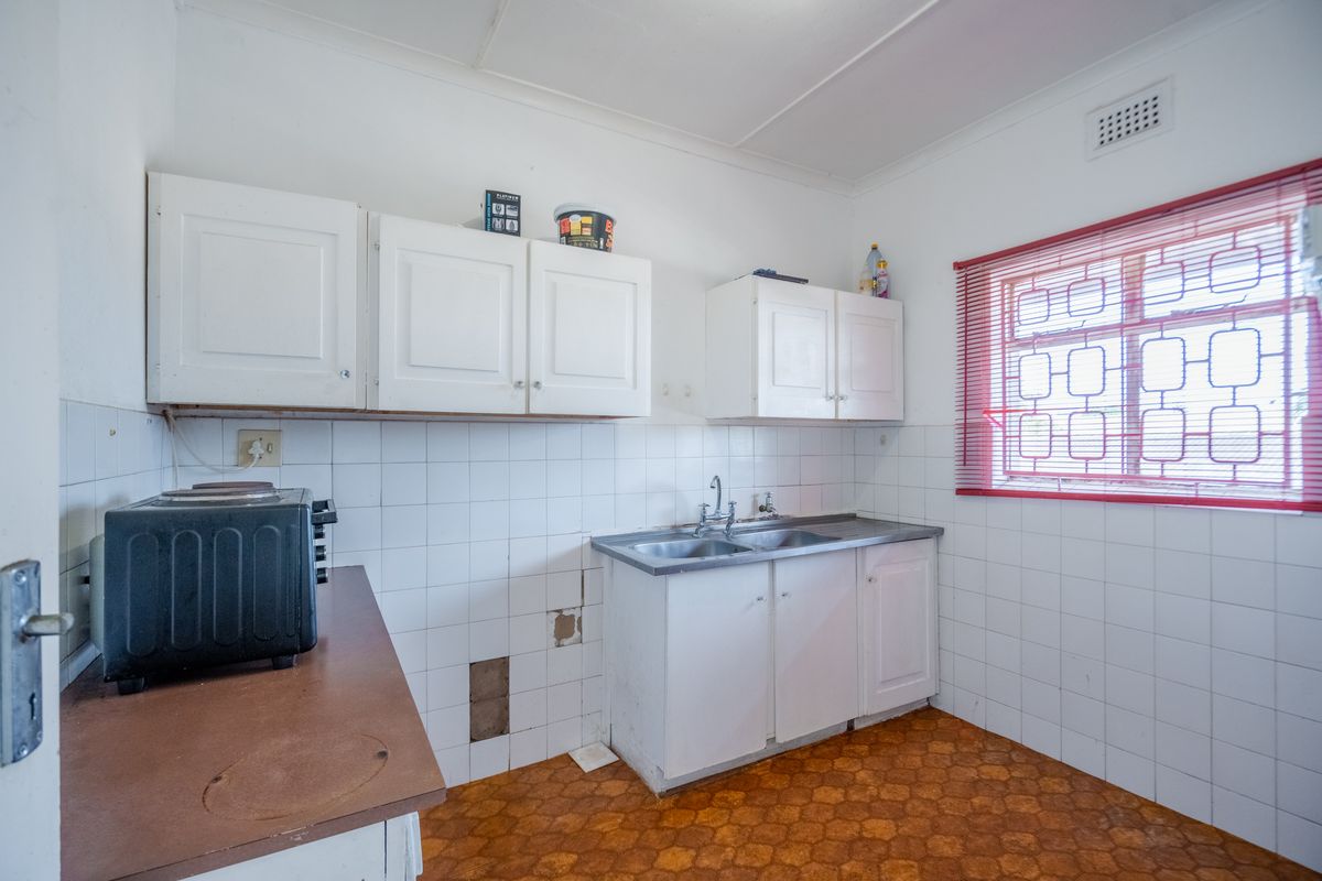 A dated kitchen featuring white cabinetry, a double bowl sink, tiled splashbacks, and a security-fitted window, requiring cosmetic updates and repairs.