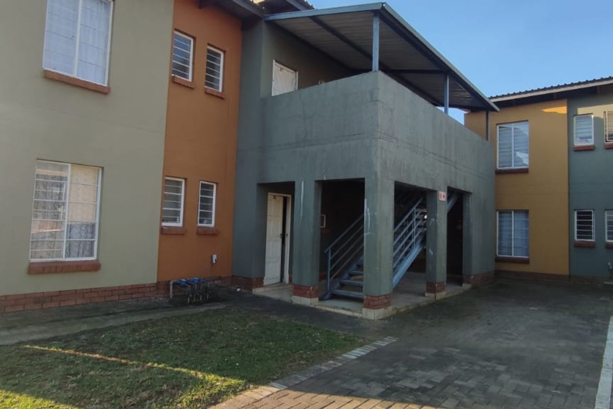 Rear view of a multi-story townhouse complex featuring paved parking, external metal staircases, and covered patio areas with painted brick and plaster finishes.