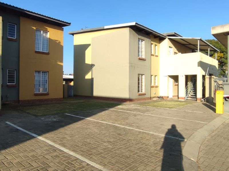 Exterior view of a townhouse complex featuring a paved driveway, security access gate, and multi-story residential units with established landscaping.