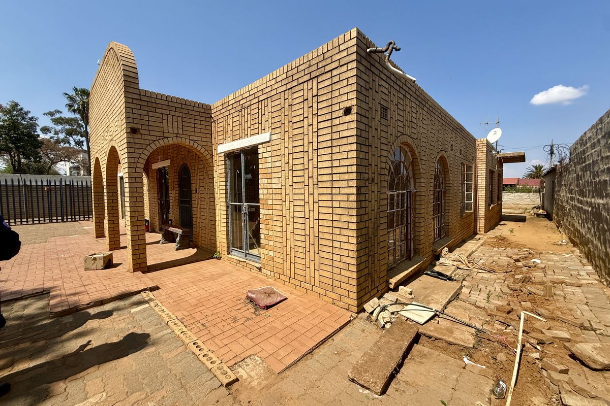 Rear view of a face brick house featuring an arched portico, paved patio area, and arched window frames, requiring exterior landscaping and maintenance.