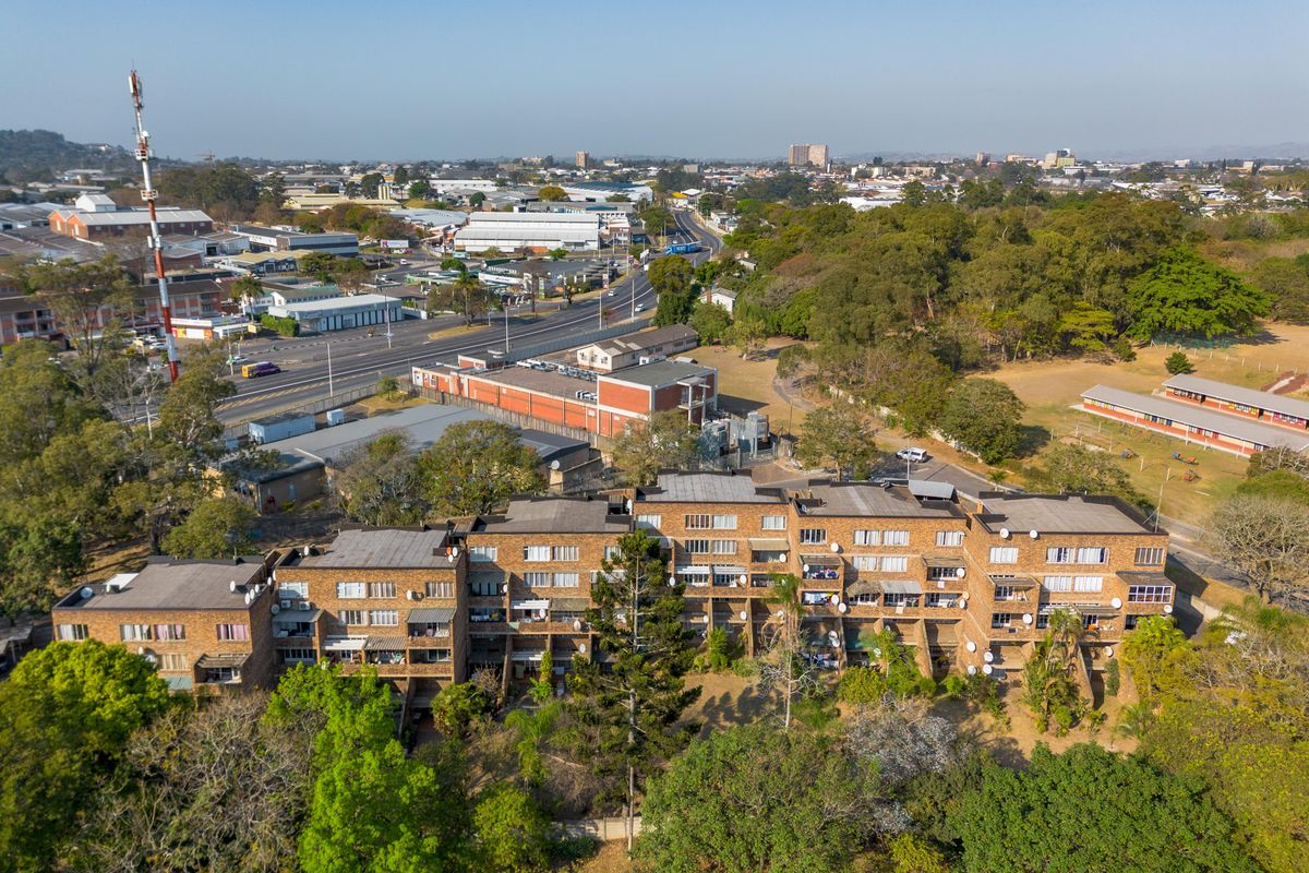 Aerial view of a multi-story brick apartment complex situated in a suburban area with surrounding greenery and nearby road infrastructure.