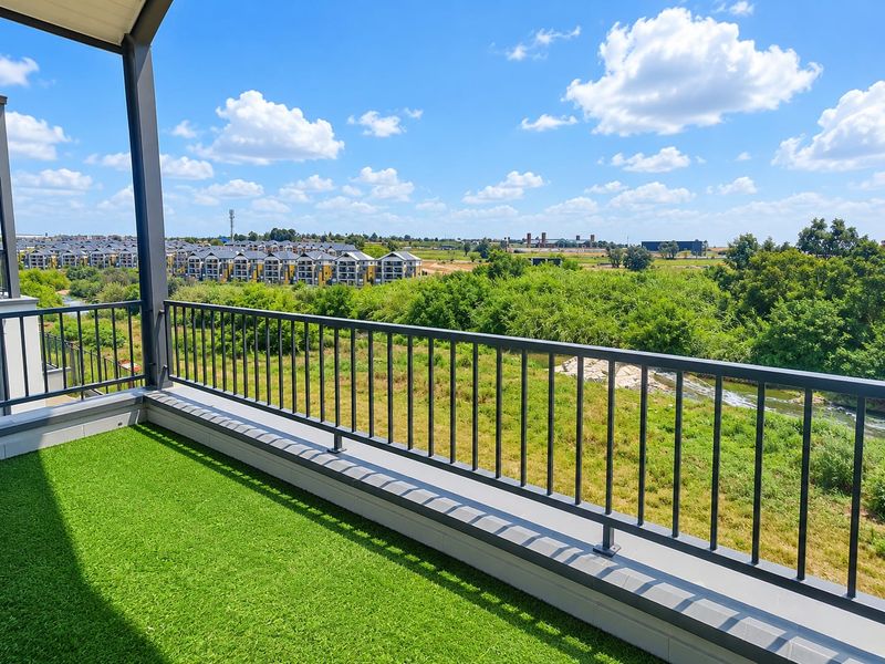A spacious balcony featuring a modern metal balustrade and artificial turf, offering expansive views of the surrounding landscape and nearby residential complex.