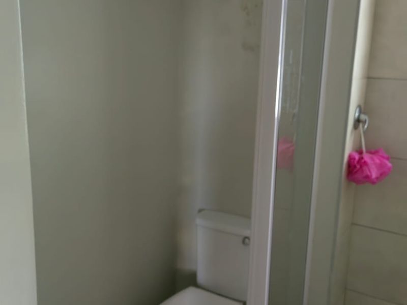 A compact bathroom featuring a toilet and a glass-enclosed shower with tiled walls, showing signs of wear and wall surface damage.