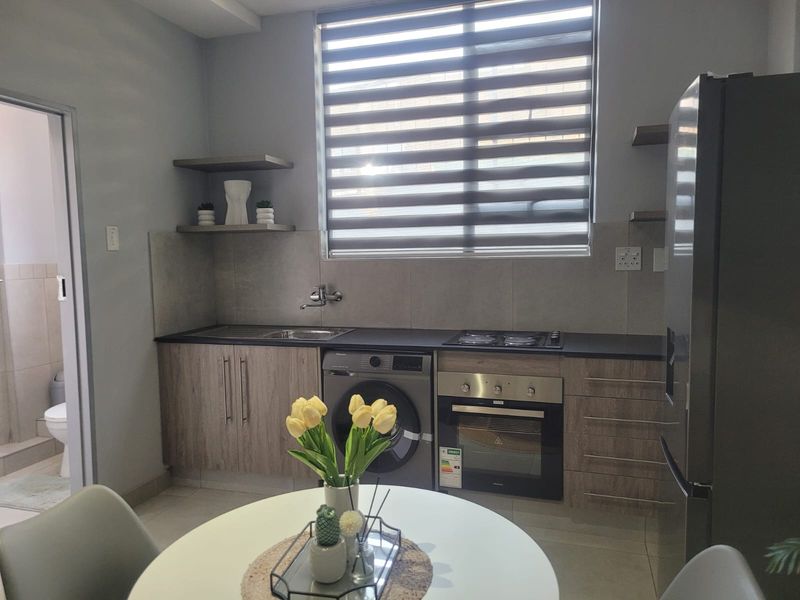 Modern kitchen featuring wood-finish cabinetry, built-in oven and hob, tiled backsplash, and integrated laundry space, complemented by a large window with roller blinds.