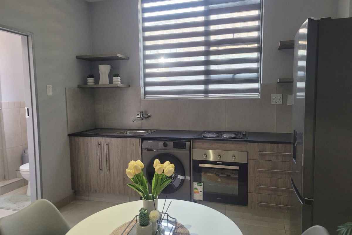 Modern kitchen featuring wood-finish cabinetry, built-in oven and hob, tiled backsplash, and integrated laundry space, complemented by a large window with roller blinds.