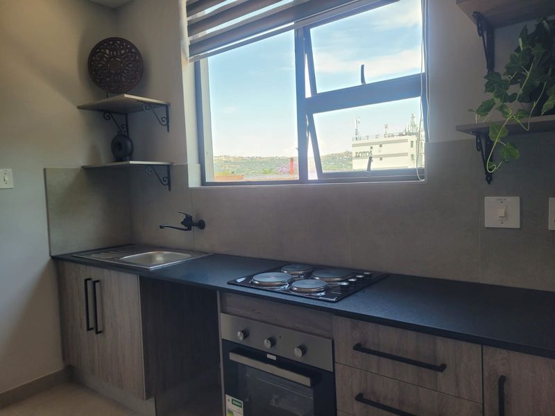 Modern kitchen featuring wood-grain cabinetry, dark laminate countertops, integrated electric hob and oven, and a functional stainless steel sink with a view.