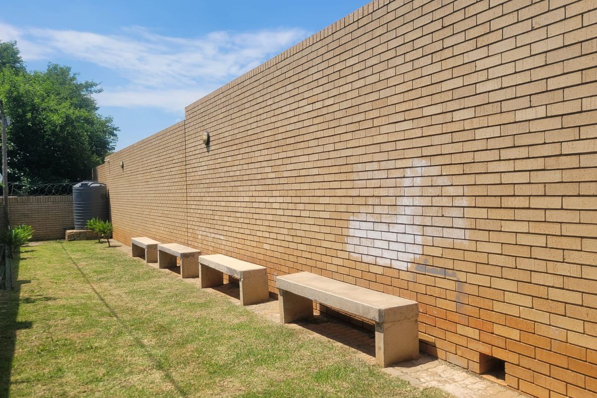 A rear exterior area featuring a long brick boundary wall, integrated concrete bench seating, and a water tank set against a lawn.