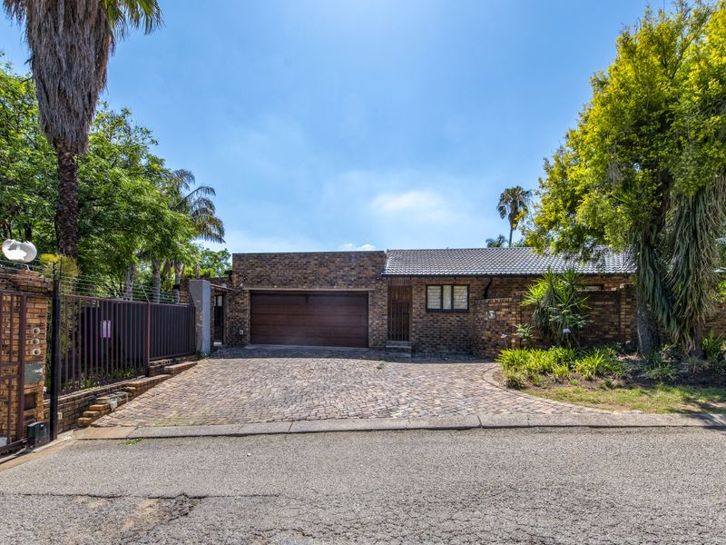 Front exterior view of a single-story brick house featuring a double garage, paved driveway, and established garden with security fencing.