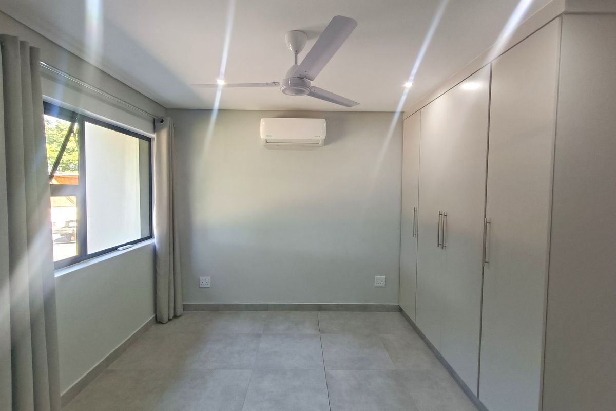 A clean, modern bedroom featuring neutral tiled flooring, built-in cupboards, a ceiling fan, and a wall-mounted air conditioning unit.