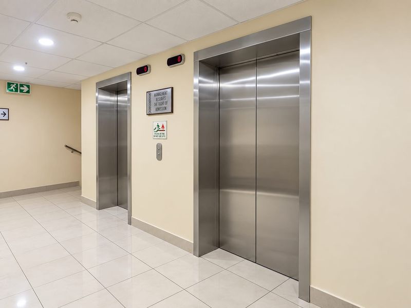 A clean and well-maintained apartment building hallway featuring stainless steel elevator doors, tiled flooring, and recessed lighting.