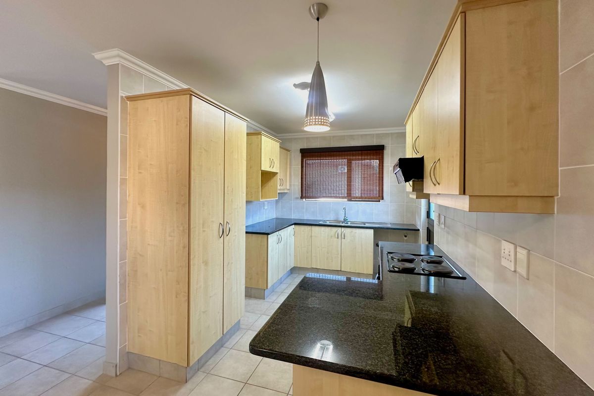 A clean, functional kitchen featuring light wood-finish cabinetry, dark granite countertops, an integrated electric hob, and neutral floor tiling.