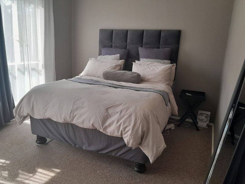 A comfortable bedroom featuring neutral wall tones, carpeted flooring, and natural light provided by a large window with curtains.