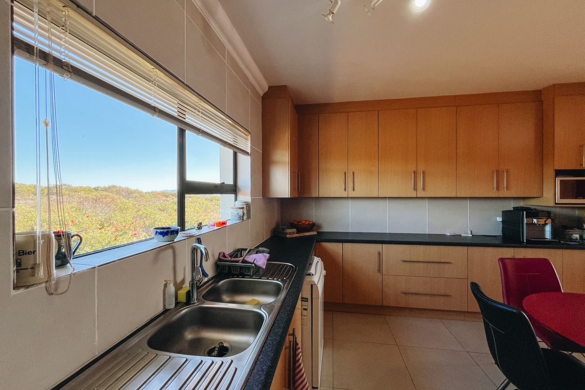 A functional kitchen featuring light wood cabinetry, dark laminate countertops, a double stainless steel sink, and large windows offering scenic natural views.