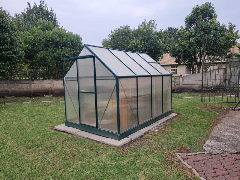 A freestanding greenhouse structure located in the rear garden, featuring a metal frame and translucent polycarbonate panels on a paved base.