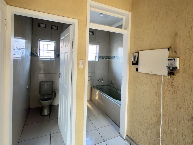A hallway area providing access to two separate bathroom spaces, featuring tiled flooring and a wall-mounted prepaid electricity meter.
