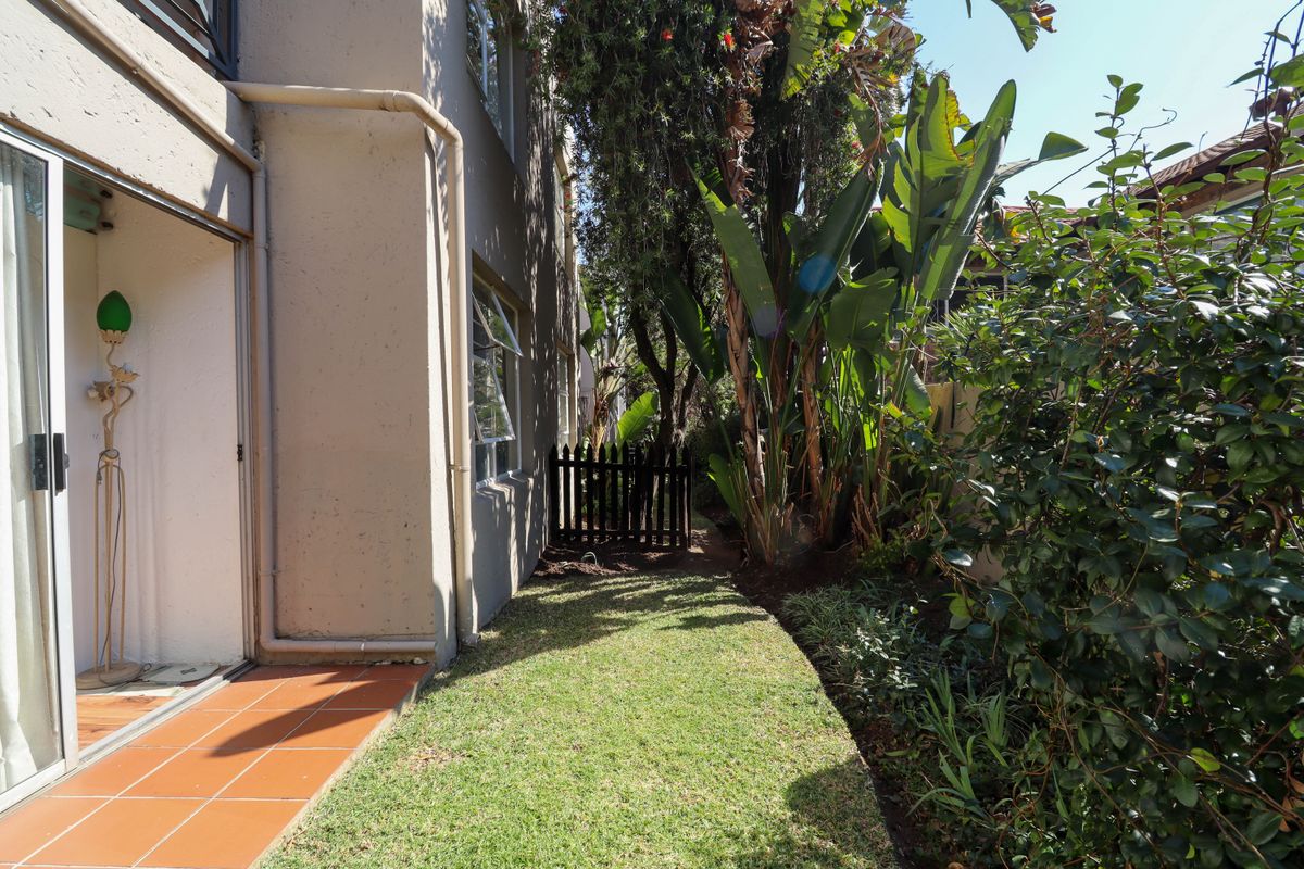 A private side garden area featuring a tiled patio, lush greenery, and a small wooden gate, providing a quiet outdoor space for the apartment.